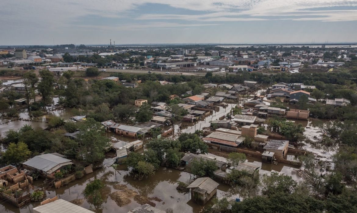 Vista aérea de Eldorado do Sul em maio de 2024: casas de um bairro ainda cercadas pela água barrenta em primeiro plano e, ao fundo, a avenida e o comércio da cidade em terreno seco.