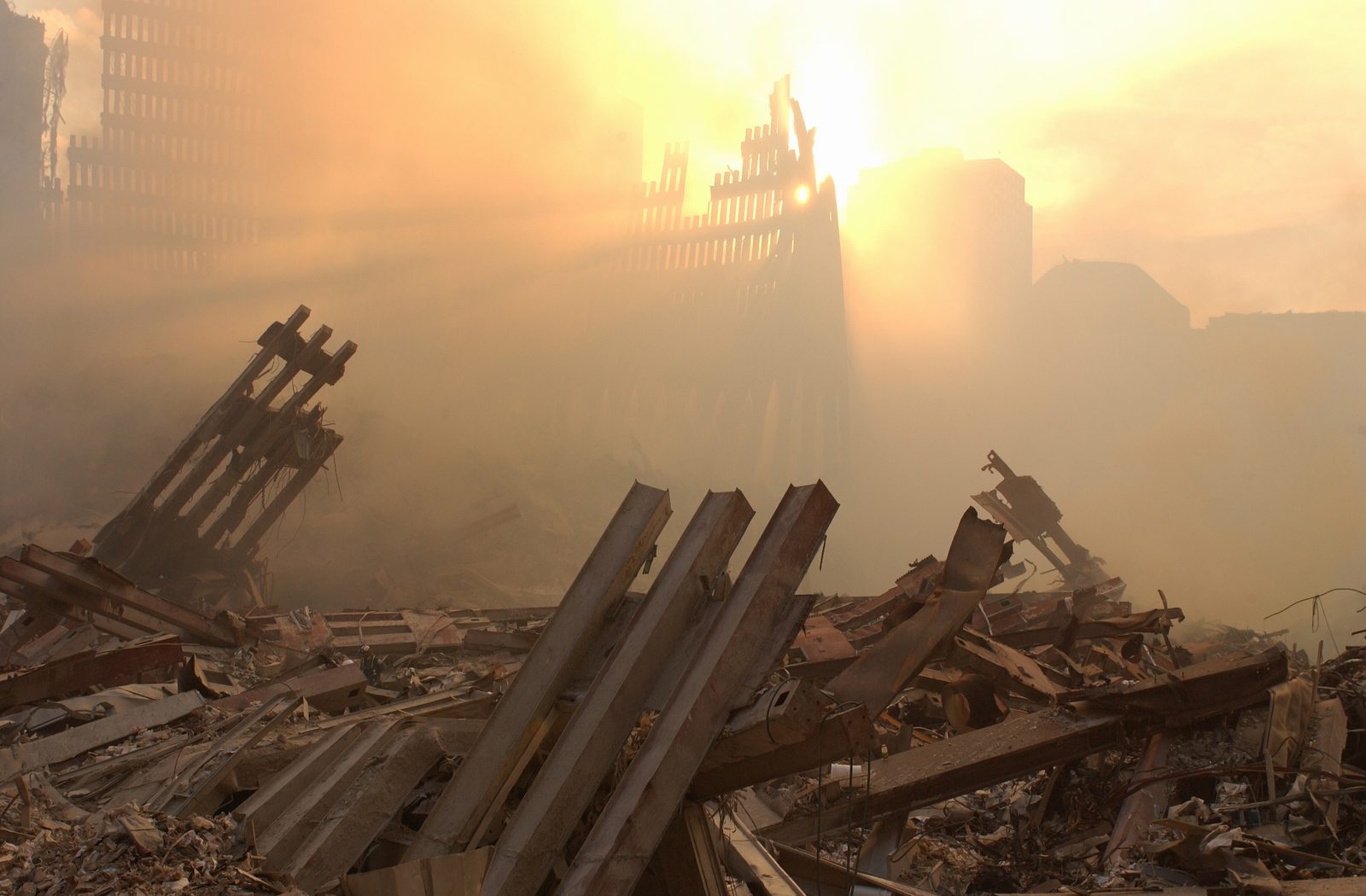 Ground Zero em Nova York nos dias seguintes ao 11 de setembro de 2001: vigas de aço retorcidas em primeiro plano e o sol atravessando a nuvem de poeira sobre os restos da fachada do World Trade Center.